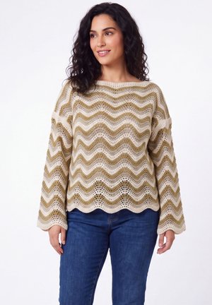 Vrouw met krullend donker haar die een beige en olijfgroene golvende gebreide trui en blauwe spijkerbroek draagt, glimlachend en naar de zijkant kijkend.
