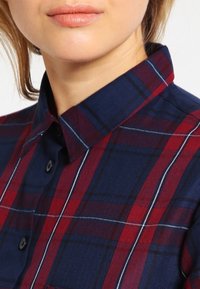 Chemise à boutons en bleu marine avec un motif en tartan rouge et blanc. Présente un col structuré et des boutons noirs, fabriquée en tissu tissé.
