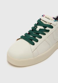 Baskets en cuir blanc avec des lacets verts, bout arrondi et semelle arrondie. Doublure à motif floral visible près de la languette. Étiquette de la marque : ALDO.