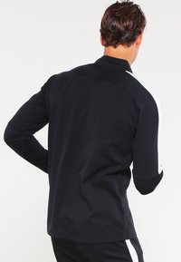 Maglione atletico nero a maniche lunghe con colletto alto, caratterizzato da strisce bianche sui lati. Materiale morbido e liscio con un design aderente.