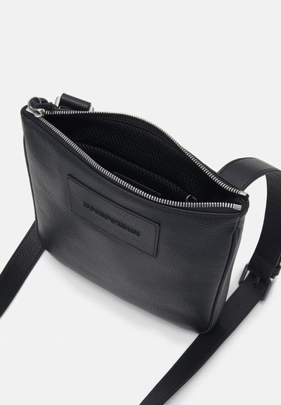 Černá kožená crossbody taška s texturovaným povrchem, zipovým uzavíráním, síťovými vnitřními přihrádkami a vyraženou logo nášivkou na přední straně.
