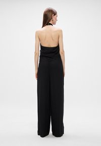 Μαύρο φόρεμα ολόσωμου στυλ με λαιμόκοψη halter, ραμμένο μπούστο και παντελόνια με φαρδιά πόδια. Ίσια ύφανση με χαλαρή εφαρμογή. Σχεδίαση με ανοιχτή πλάτη.
