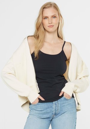 Cremfarbener Reißverschluss-Hoodie mit gerippten Bündchen über einem schwarzen Tanktop. Kombiniert mit hellblauen Jeans. Hat eine lässige Passform und eine weiche Textur.