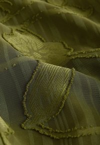 Tissu vert olive avec motifs de feuilles brodés texturés et sections rayées transparentes visibles en gros plan.