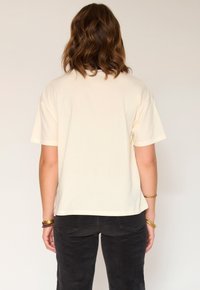 Femme aux cheveux bruns mi-longs portant une chemise beige unie à manches courtes et un pantalon noir, se tenant de dos contre un fond clair.