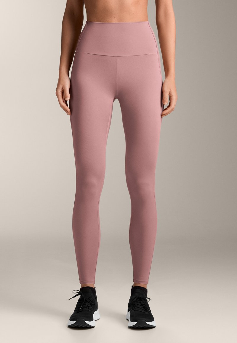 Legíny s vysokým pásom z jemnej ružovej tkaniny s hladkým povrchom, s minimalistickým dizajnom, v kombinácii s čiernymi športovými topánkami.