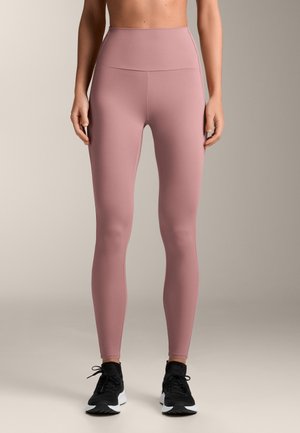 Colanți cu talie înaltă dintr-un material roz pal, cu finisaj neted, având un design minimalist și asortați cu pantofi sport negri.