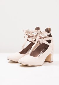 Scarpe con tacco alto in suede beige con design a T, apertura sul retro e dettagli in pizzo. Punta arrotondata e tacco a blocco di 7 cm per stabilità.