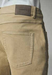 Khaki bukser med en glat tekstur, en baglomme og et mørkt lædermærke med teksten "DENIM SUPPLY Co." ved taljen.