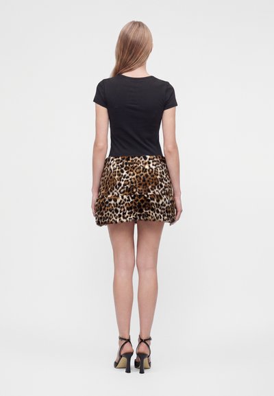 Femme debout, de dos, portant un haut noir à manches courtes, une mini-jupe imprimé léopard et des sandales noires à talons hauts sur fond blanc.