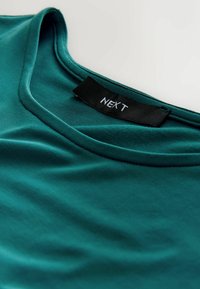 Türkisfarbenes Baumwoll-T-Shirt mit rundem Halsausschnitt und glatter Textur; verfügt über ein schwarzes Etikett mit "NEXT" in weißer Schrift.