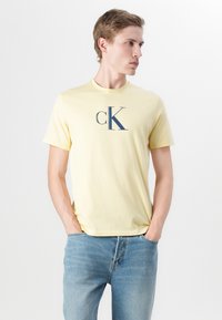 Žuta pamučna majica s okruglim izrezom, kratkim rukavima i tirkiznim "CK" logom otisnutim na prednjem dijelu, u kombinaciji sa svijetloplavim trapericama.