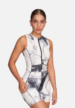 Frau trägt einen ärmellosen, vorne mit Reißverschluss ausgestatteten sportlichen Overall mit abstrakten schwarz-weißen Mustern, stehend vor einem einfarbigen hellen Hintergrund.