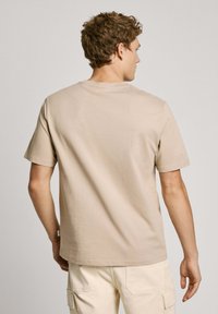Beige �īsām piedurknēm t-krekls, izgatavots no kokvilnas. Tam ir apaļa kakla izgriezums un maiga tekstūra. Aizmugures skats ar minimālu šuvju detaļām.