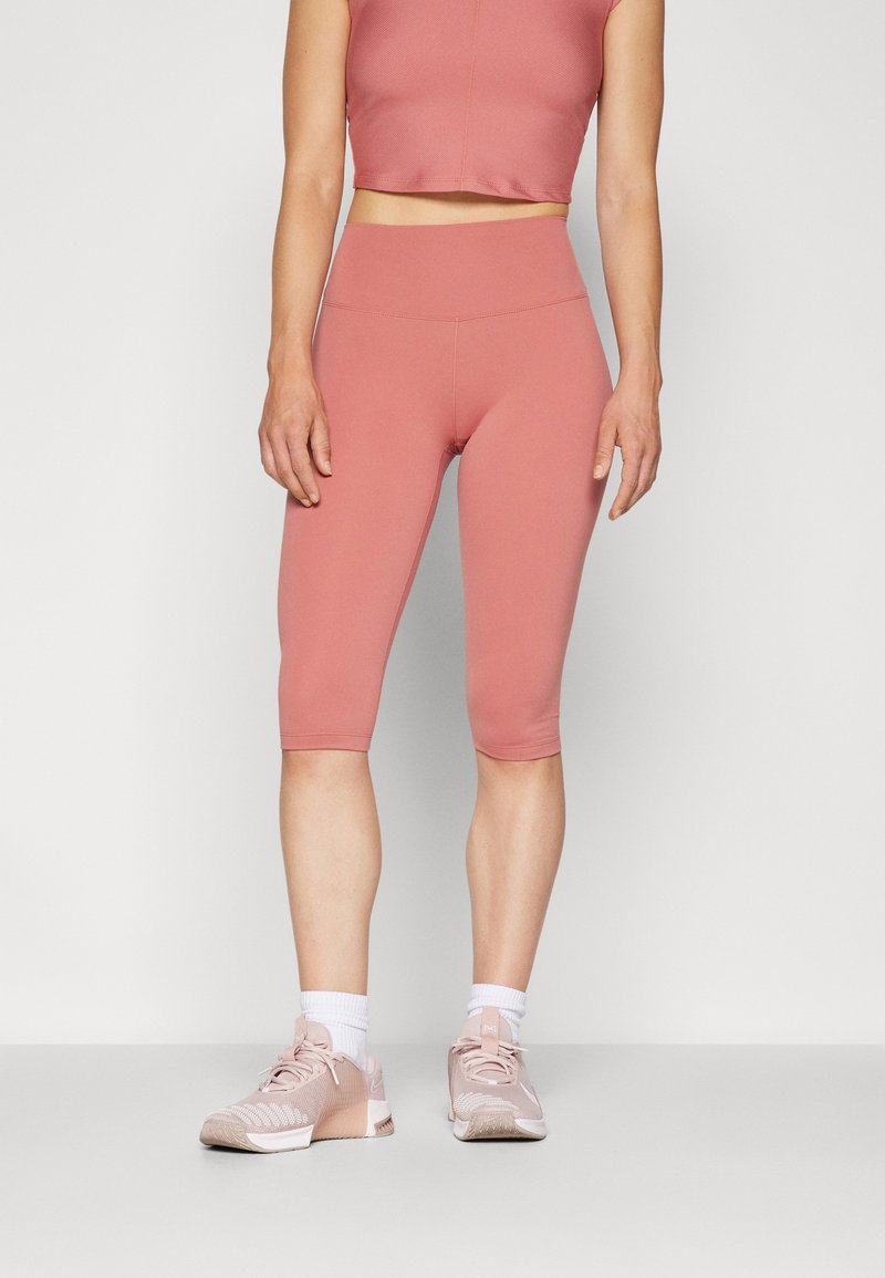 Różowy, dopasowany crop top w połączeniu z pasującymi legginsami do kolan, oba wykonane z gładkiego, elastycznego materiału. Noszony z lekkimi, różowymi trampkami.
