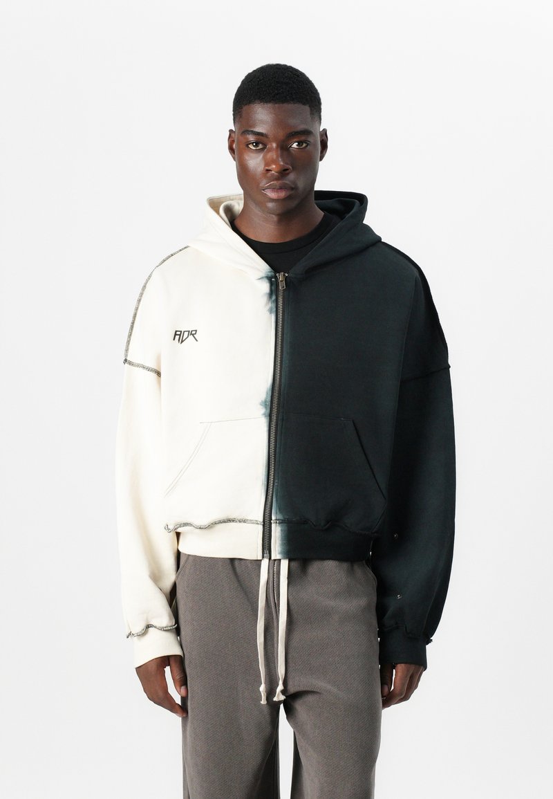 Sweatshirt com capuz e fecho éclair, com design dividido; uma metade branca, outra metade preta, com uma barra desgastada e costura em contraste. Possui um bolso frontal.