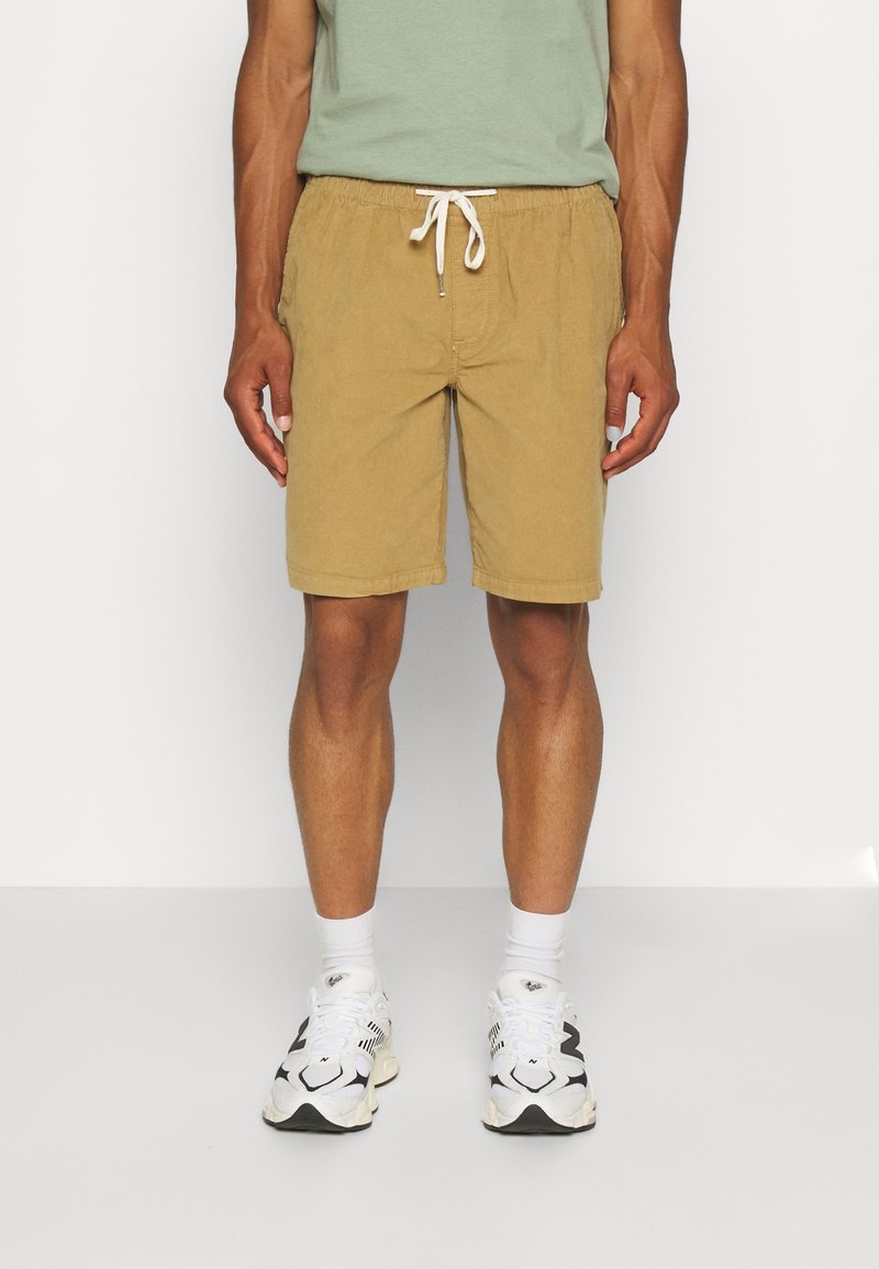 Shorts en coton kaki avec une taille élastique et un cordon de serrage, avec une coupe décontractée et des poches latérales, associés à des baskets blanches.