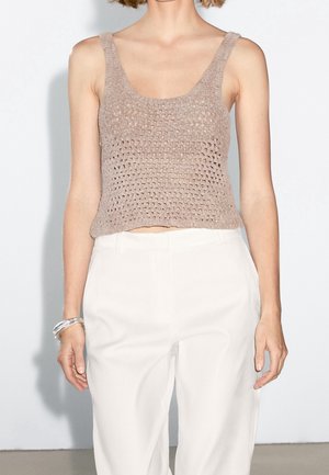 Kvinde iført beige hæklede ærmeløse croptop, hvide bukser med høj talje og en gennemsigtig armbånd på venstre håndled, stående mod en hvid væg.
