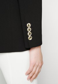 Manche de blazer noir avec quatre boutons dorés brillants et une main posée à côté d'un pantalon blanc.