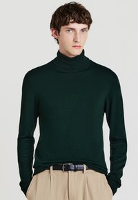 Grüner Rollkragenpullover aus glattem Material, mit langen Ärmeln und figurnaher Passform. Kombiniert mit beigen Hosen und einem schwarzen Gürtel.