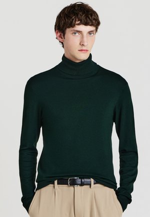 Grüner Rollkragenpullover aus glattem Material, mit langen Ärmeln und figurnaher Passform. Kombiniert mit beigen Hosen und einem schwarzen Gürtel.