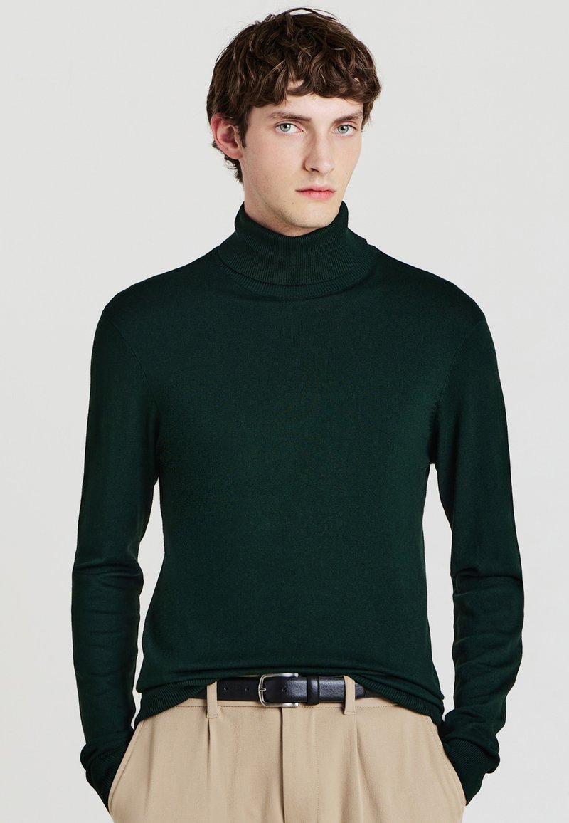Grüner Rollkragenpullover aus glattem Material, mit langen Ärmeln und figurnaher Passform. Kombiniert mit beigen Hosen und einem schwarzen Gürtel.