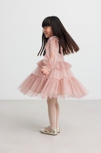 Ein lächelndes Mädchen trägt ein geschichtetes pinkes Kleid mit Rüschen und goldenen Ballettschuhen, während es sich vor einem schlichten weißen Hintergrund dreht.
