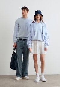 Άνδρας με φωτεινό γαλάζιο πουλόβερ και τζιν κρατώντας μαύρη τσάντα, γυναίκα με φωτεινό γαλάζιο μπουφάν, πλισέ λευκή φούστα, λευκά αθλητικά παπούτσια και σκούρο μπλε καπέλο τύπου κάλπη.