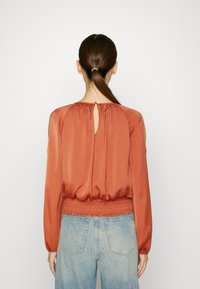 Blusa de cor rust com mangas longas, bainha elástica e detalhe de abertura nas costas. Combinada com jeans de cintura alta de cor azul clara. Textura suave.
