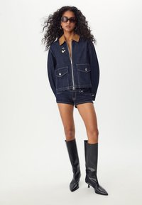Veste en denim avec un col en velours côtelé marron, deux poches avant et quincaillerie argentée. Associée à un short en denim et des bottes noires jusqu'aux genoux.