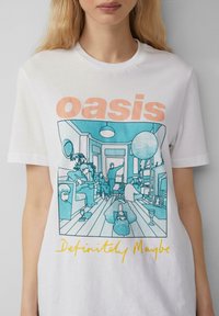 T-shirt blanc arborant un graphique bleu représentant une scène de salon avec des personnages, des guitares, un grand ballon et un texte indiquant « oasis Definitely Maybe ». 