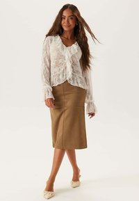 Blouse en dentelle avec un col et des manchettes à volants, associée à une jupe midi brune ajustée. Le look est complété par des chaussures de couleur claire.