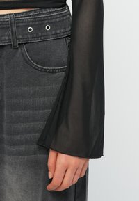 Κοντινό πλάνο ενός χεριού δίπλα σε ένα μαύρο διάφανο πουκάμισο με μακριά μανίκια και σκούρα γκρι τζιν ψηλόμεσα με φαρδιά ζώνη και ασημί κουμπώματα.