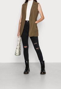 Brun ärmlös blazer, vit ribbad topp, svarta slitna skinny jeans, svarta snörboots och en vit väskhandväska i ormskinn.