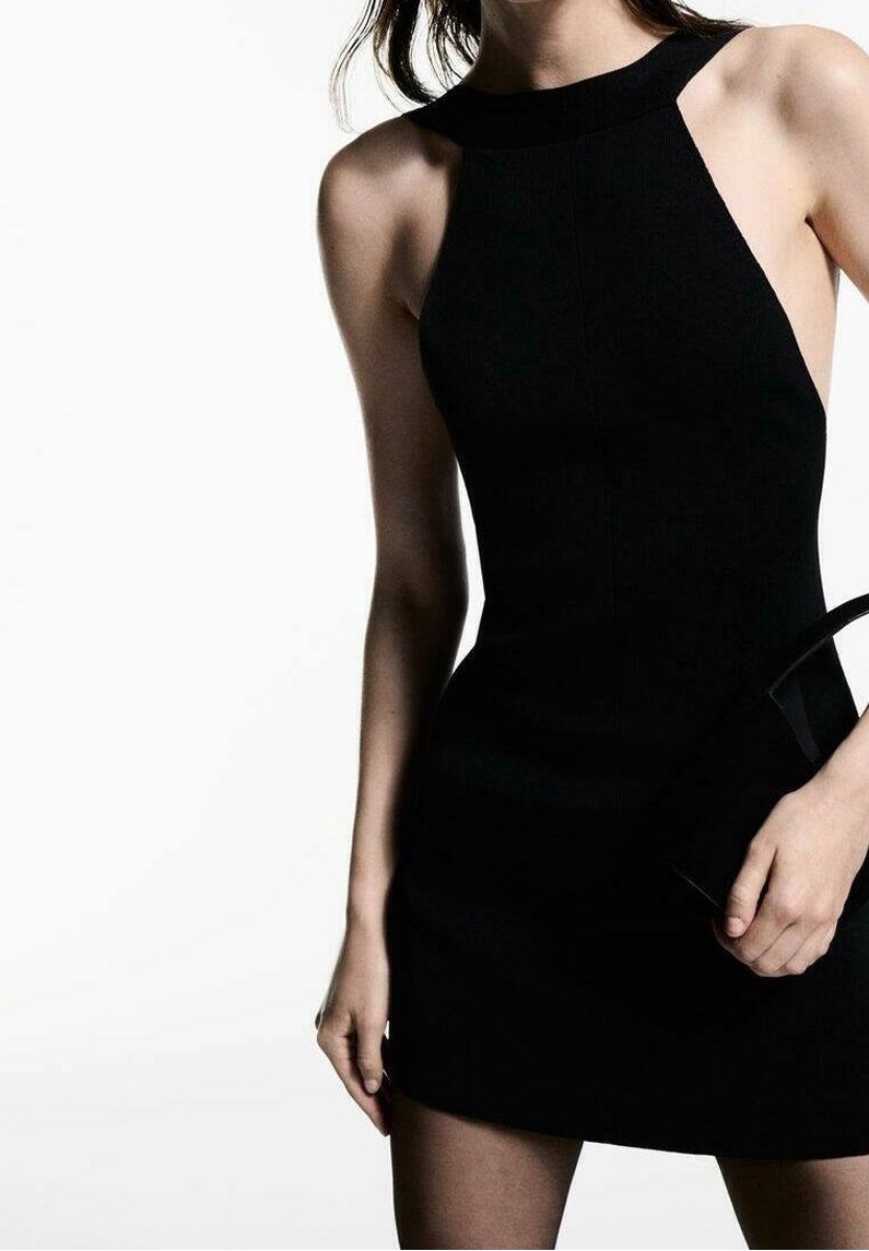 Robe noire sans manches avec un col montant et une silhouette ajustée, confectionnée dans un matériau texturé. Petit sac à main assorti à la main.