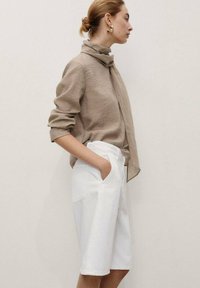 Une femme se tient de profil, portant un chemisier beige et un short blanc, avec les cheveux en chignon et une écharpe autour du cou, devant un arrière-plan neutre.