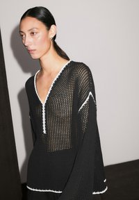 Femme aux cheveux foncés en queue de cheval basse, portant un pull noir à mailles lâches avec des détails blancs et un col en V, se tenant devant un fond uni.