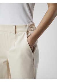 Beige housut, joissa on ribboitettu joustavyö, sivutaskut ja muotoiltu malli. Kangas vaikuttaa pehmeältä ja sileältä.