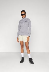 Grijze fleece jack met hoge kraag en ritszak. Gepaarde met lichtbeige shorts en zwarte wandelschoenen. Witte sokken.
