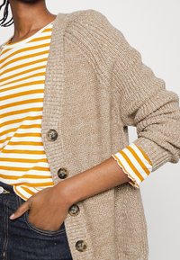 Beige stickad cardigan med ribbad textur, utrustad med stora bruna knappar och en avslappnad passform. Underneath bärs en gul och vit randig skjorta med volangärmar.