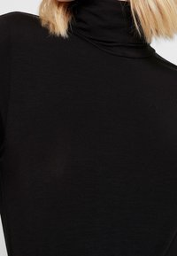 Maglietta nera a collo alto con un design aderente, caratterizzata da una texture liscia e una leggera costolatura. Il tessuto appare elastico e morbido.