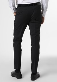 Man med svarta kostymbyxor, svart bälte, svarta kostymskor och vit långärmad skjorta som står med händerna i fickorna, sett bakifrån.