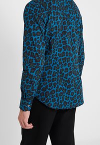 Blauwe luipaardprint blouse met lange mouwen, met een zwarte achtergrond en een gebogen zoom. Inclusief opgesloten manchetten en een gladde textuur.