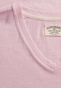 T-shirt en coton rose clair à col en V avec une étiquette en tissu indiquant "Superdry Co. Craft & Detail British Design" à l'intérieur du col.
