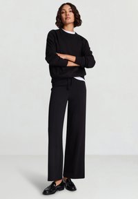 Schwarzer Pullover mit rundem Ausschnitt und langen Ärmeln, kombiniert mit schwarzen Hosen mit weitem Bein. Gestylt mit schwarzen Loafers.