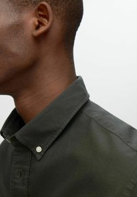 Camisa verde oscuro de botones con cuello puntiagudo, que muestra una sutil textura y un solo botón de perla en el cuello. Retrato con gran acercamiento.