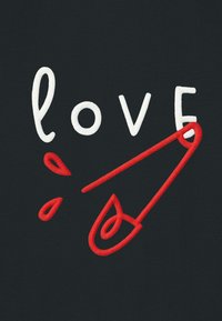 Lettere ricamate bianche che formano la parola "love" con una spilla di sicurezza rossa al posto della lettera "v" su uno sfondo di tessuto nero.