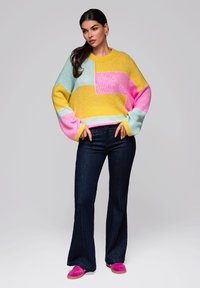 Kleurvolle gebreide trui met gele, roze en blauwe secties, gecombineerd met donkere flared jeans en felle roze sneakers.