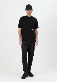 Črna majica s kratkimi rokavi z majhnim belim logotipom spredaj, v kombinaciji s hlačami v črno-belo houndstooth vzoru in črnimi supergami.