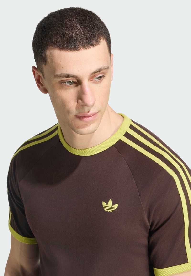 Junger Mann mit kurzen Haaren, der ein dunkelbraunes T-Shirt mit hellgrünen Streifen an den Ärmeln und einem kleinen Logo auf der Brust trägt, schaut seitlich.